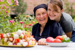 A young woman - grandchild or carer - next to an old woman at the table, with fresh food in front of them.