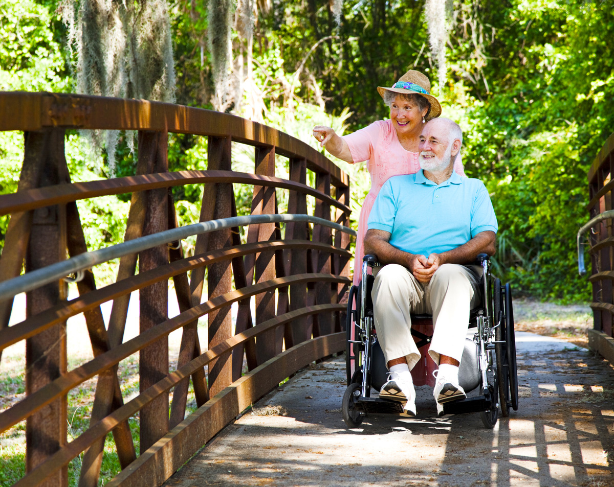 Senior woman pushing her disabled husband through the park in his wheelchair.